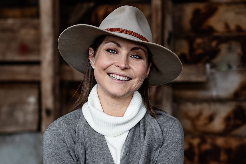 A smiling woman in a felt hat and jumper in a shed. 