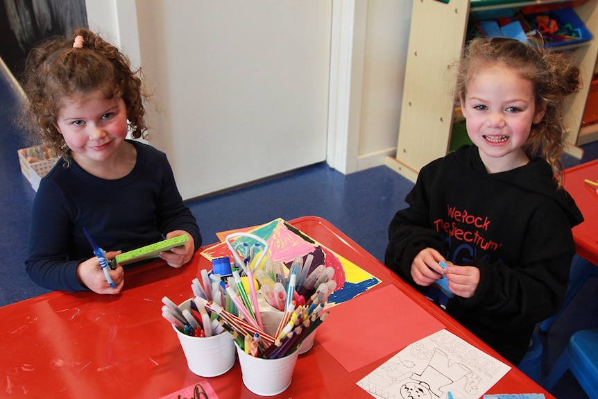 Two young girls sitting at a red table with coloured pencils and paper.