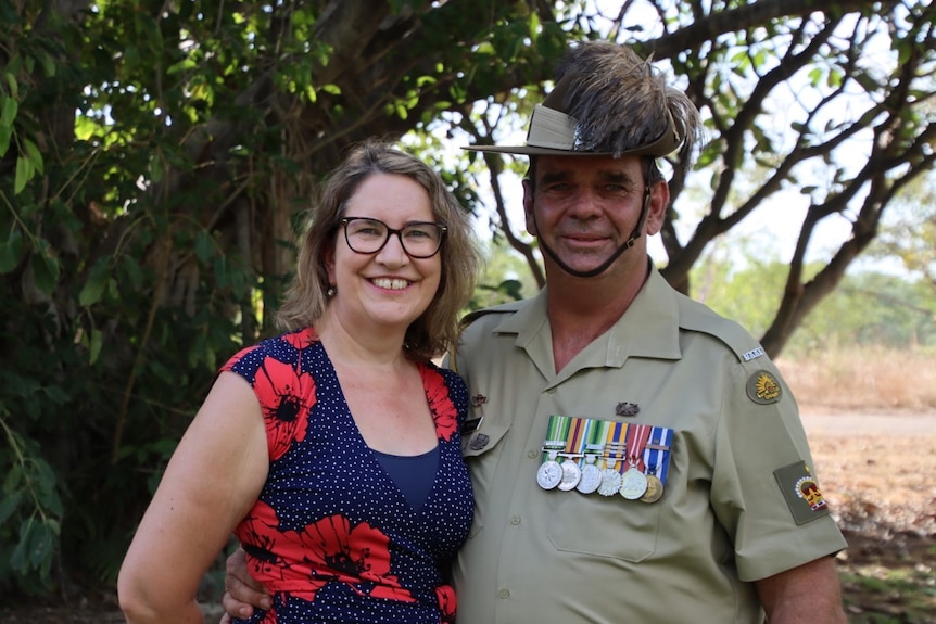 A woman in a navy dress with red flowers and a man in military uniform with medals smile together.