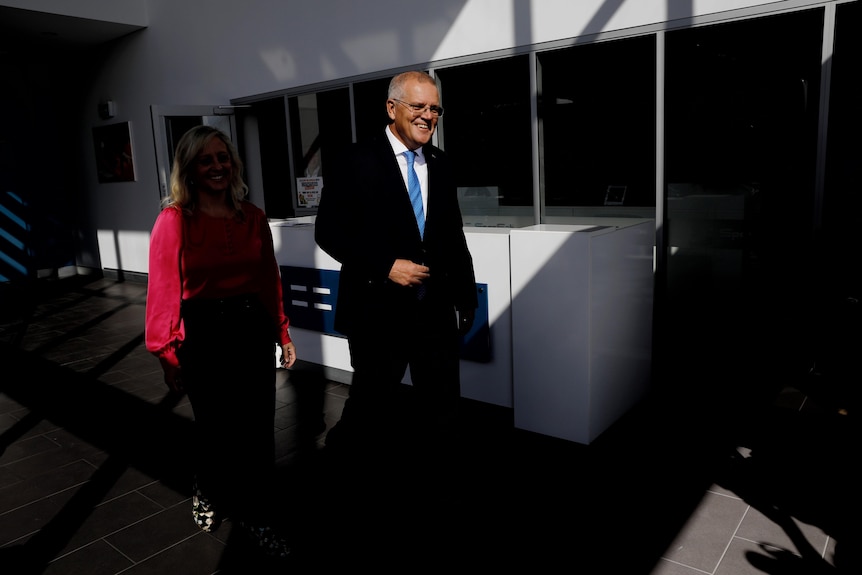 Scott Morrison wearing a suit walks outside in the sunlight next to a woman who is in the shadows. 