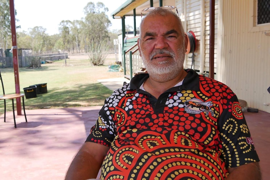 A man, wearing a bright shirt with Aboriginal artworks, sits on a patio with bushland in the background.