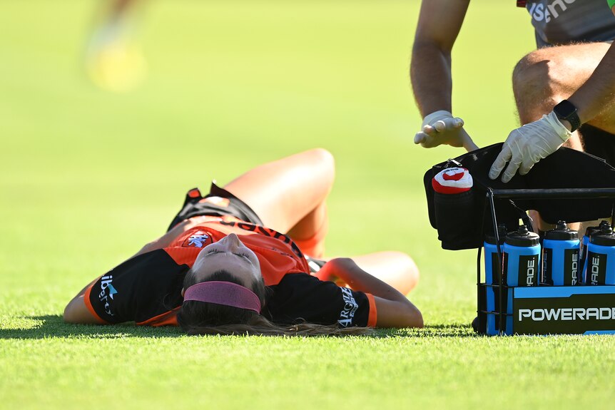 A soccer player wearing orange and black lays on her back in the grass as a doctor wearing rubber gloves attends to her