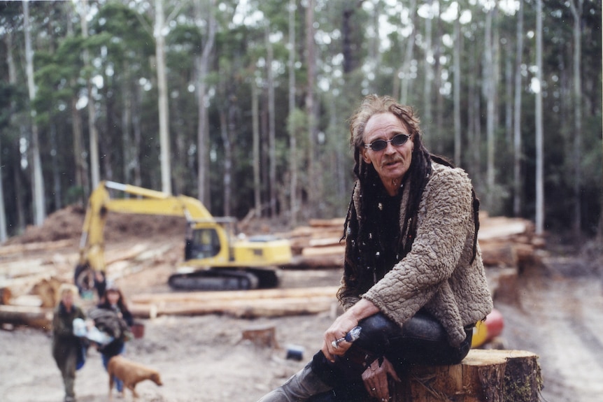 A man wearing alternative clothing sits on a tree stump, logging machinery and sawn logs in the background.