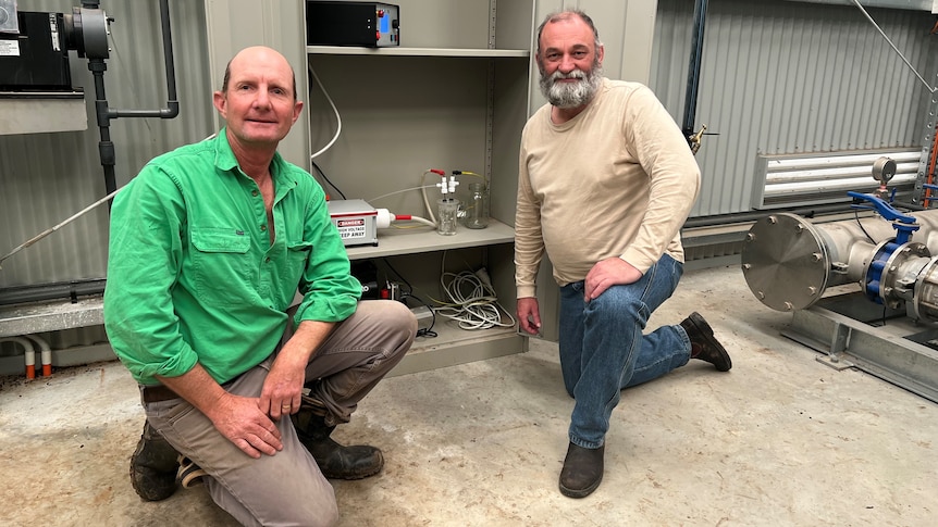 Two men kneel beside a small air compressor forcing water into a beaker with a light in the water