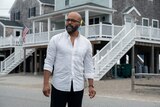 Jeffrey Wright as Monk wearing a white buttoned shirt, standing in front of a property with an American flag in the distance