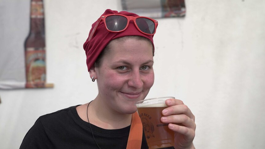 A Caucasian woman with a red head covering smiles as she holds up a cup of beer near her mouth