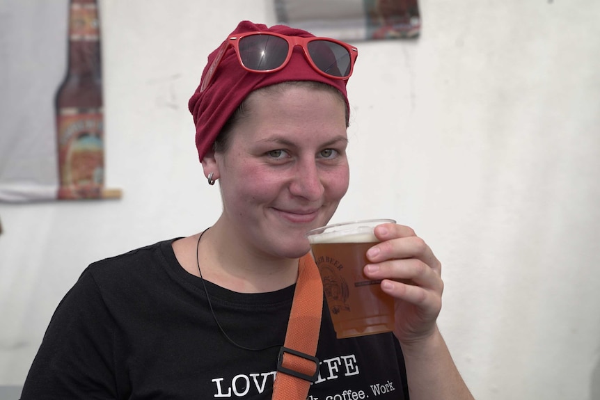 A Caucasian woman with a red head covering smiles as she holds up a cup of beer near her mouth