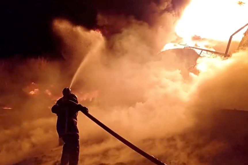 A firefighter in silhoutte sprays a hose on a large fire at night.