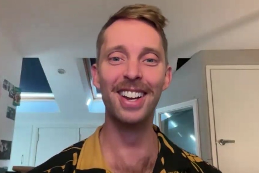 A man with dark blonde hair and a moustache smiles into a webcam. He has a yellow and black shirt.