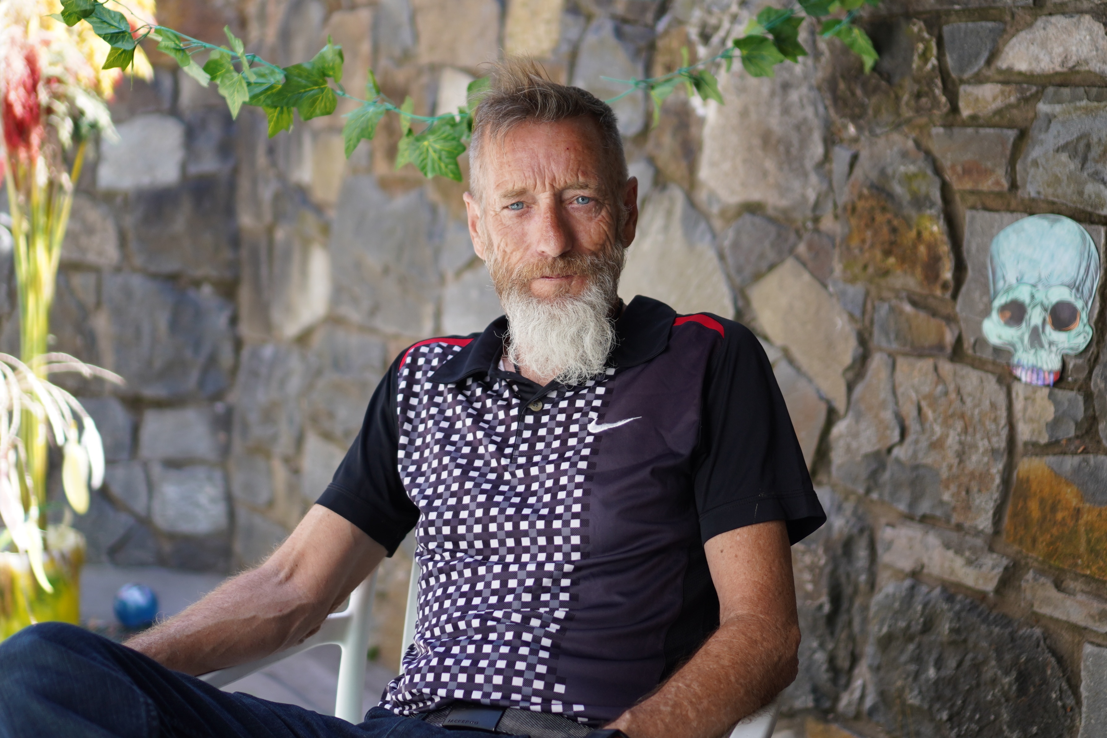 A man with a grey beard sits at an outdoor table, smiling at the camera.