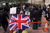 A silver-haired woman in black facemask holds UK flag in front of crowd of black-clothed protesters.