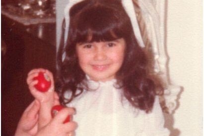 A young girl with a white ribbon in her hair holds a red Easter egg