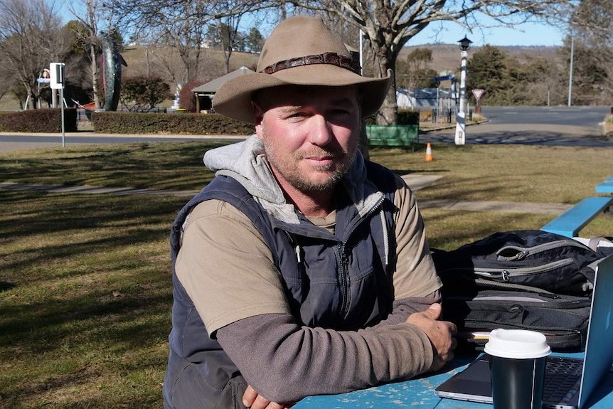 A young man wearing an Akubra, sitting at a table in a park with a coffee and his laptop.