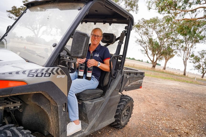 A woman wearing jeans and a polo top sits in a quad bike holding two bottles of wine, smiling.