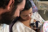 A newborn baby in his mother's arms with father watching in a hospital, in a story about supporting parents through infant loss