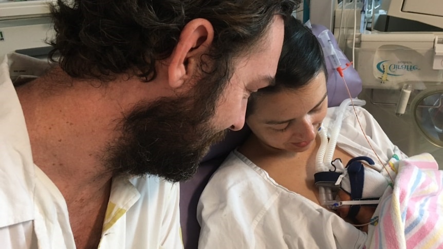 A newborn baby in his mother's arms with father watching in a hospital, in a story about supporting parents through infant loss