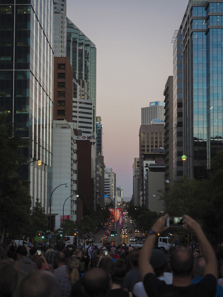 A view down St Georges Terrace at dusk showing crowds looking up at the sky, many holding smartphones up.