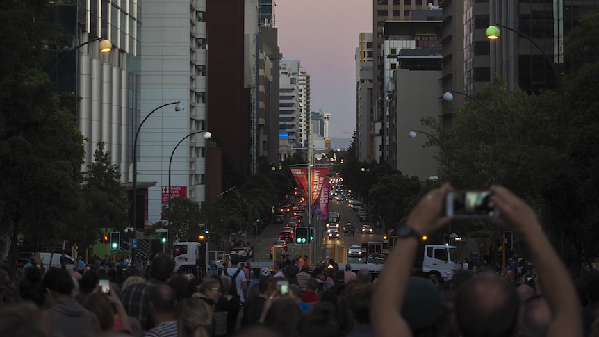 A view down St Georges Terrace at dusk showing crowds looking up at the sky, many holding smartphones up.