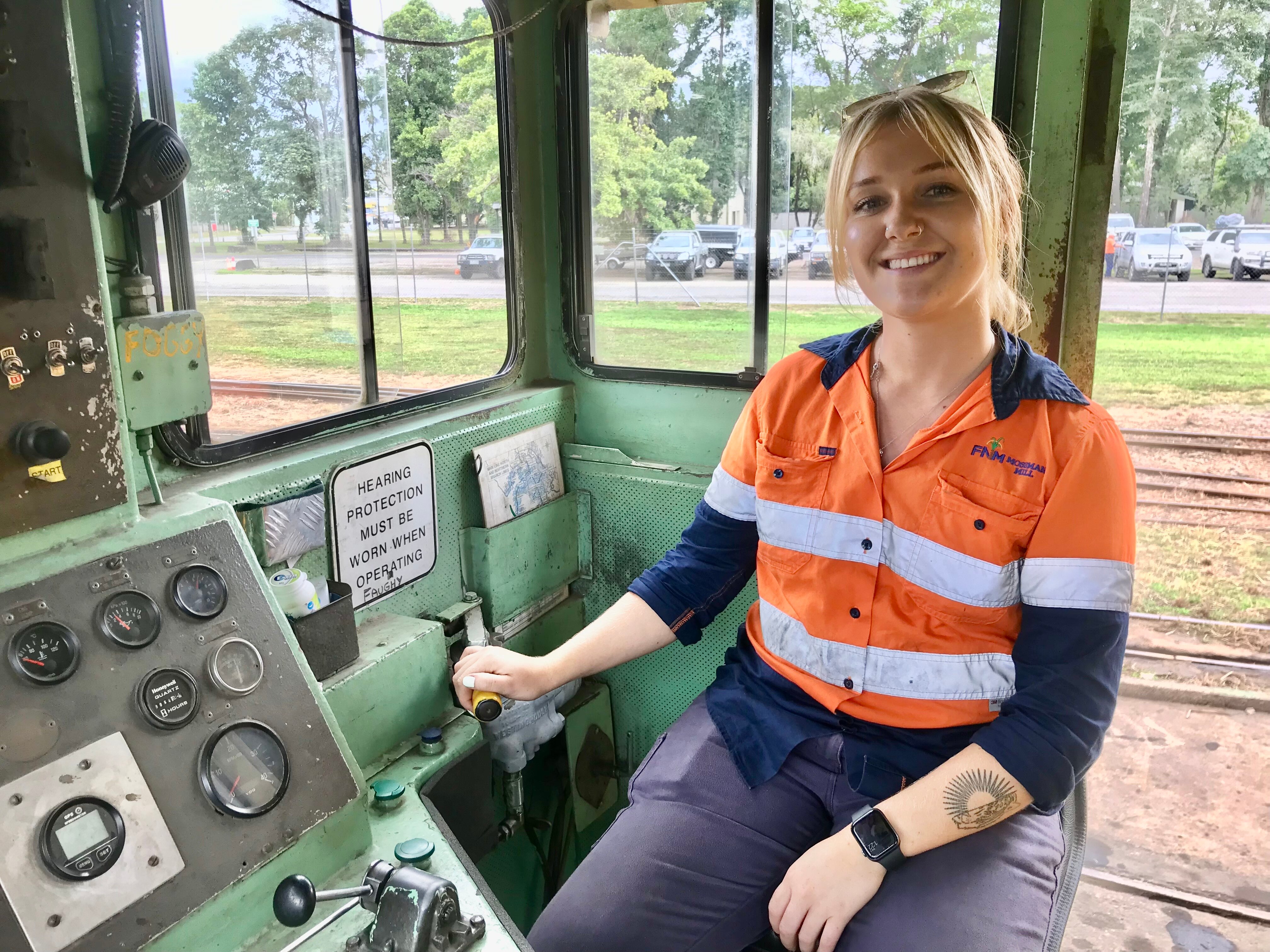 A young woman with a blonde ponytail and high-vis workwear sits in the cab of a cane train.