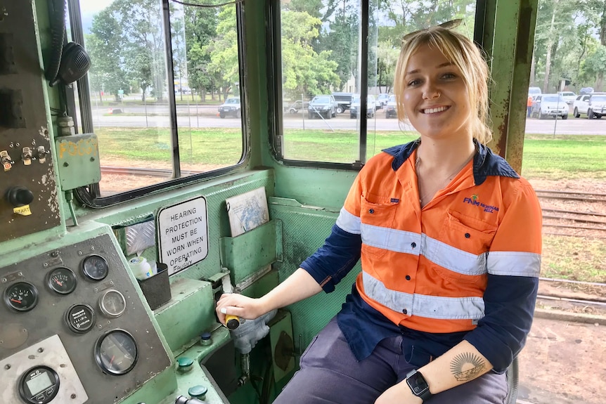 A young woman with a blonde ponytail and high-vis workwear sits in the cab of a cane train.