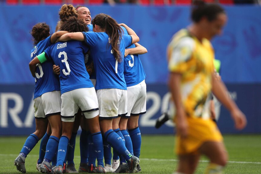 Italian players celebrate in a huddle as an Australian player walks with her head down in the foreground.