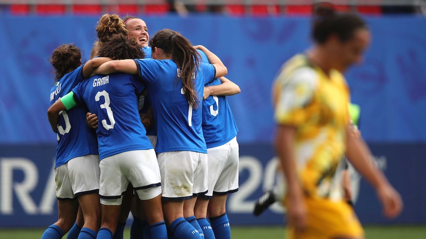 Italian players celebrate in a huddle as an Australian player walks with her head down in the foreground.