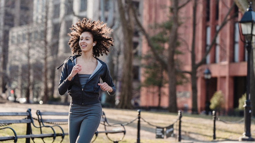 Woman jogging otuside wearing wireless earbuds with park visible in background