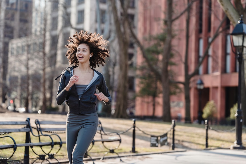Woman jogging otuside wearing wireless earbuds with park visible in background