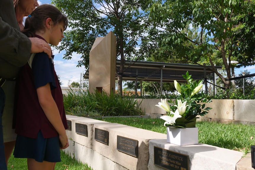 Girl in school uniform stands in front of memorial plaque with flowers on it. 
