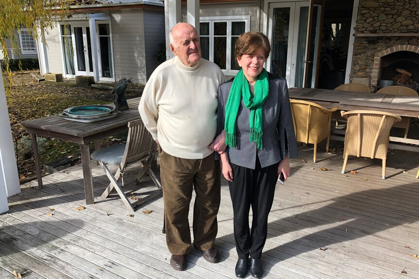 A bald man in his 90s in a white turtleneck (the artist John Olsen) and a woman in her 60s standing on a deck outside