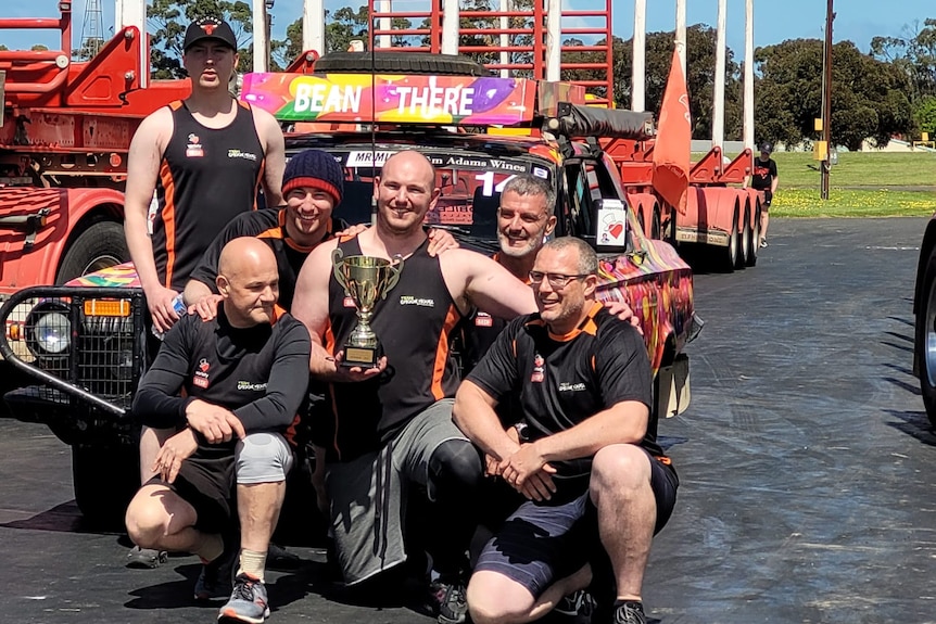 6 men with a trophy sitting on the front of a car 