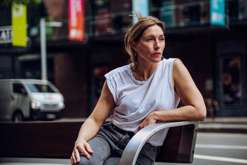 Woman in a white T shirt and jeans sits on a bench on a city street looking at to the distance on her right 