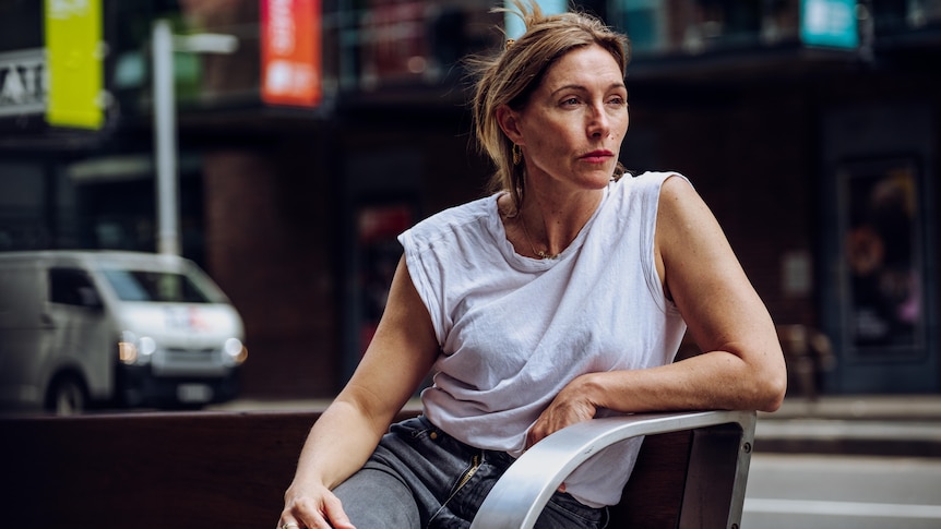 Claudia Karvan in white T-shirt and jeans sits on a bench on a city street looking to her right 