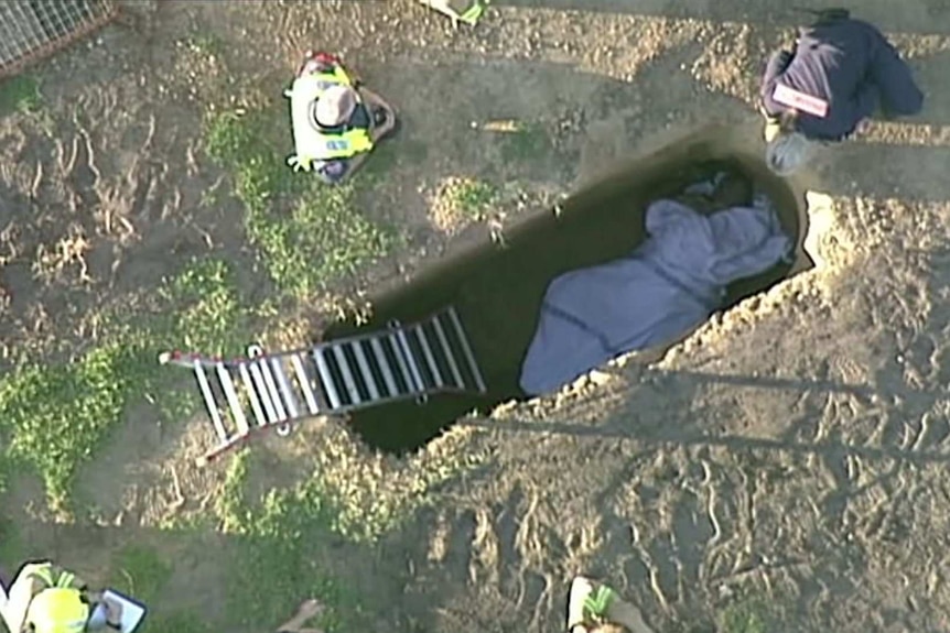 An aerial shot of a man at the bottom of a grave wrapped in a blanket with emergency workers above.