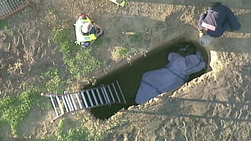 An aerial shot of a man at the bottom of a grave wrapped in a blanket with emergency workers above.