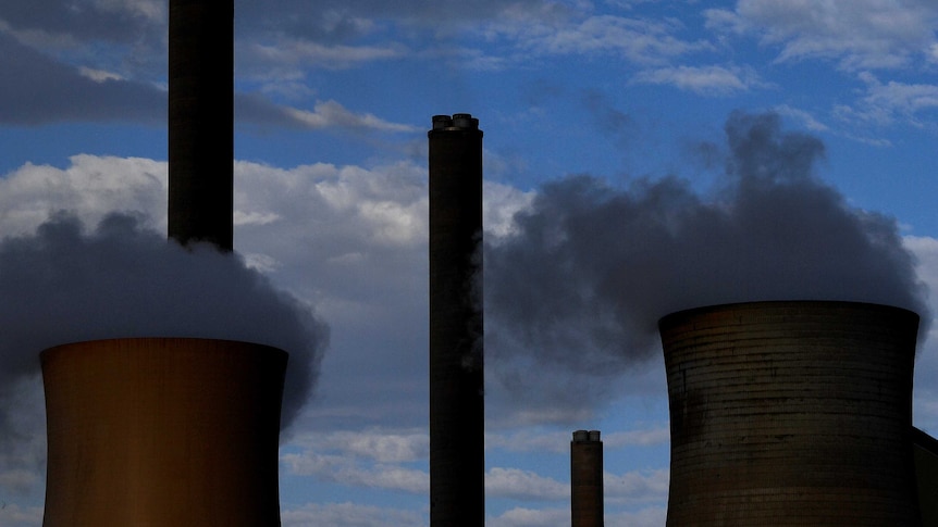 Smoke billows from two towers of a power plant as clouds obscure blue sky in the background.