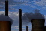 Smoke billows from two towers of a power plant as clouds obscure blue sky in the background.
