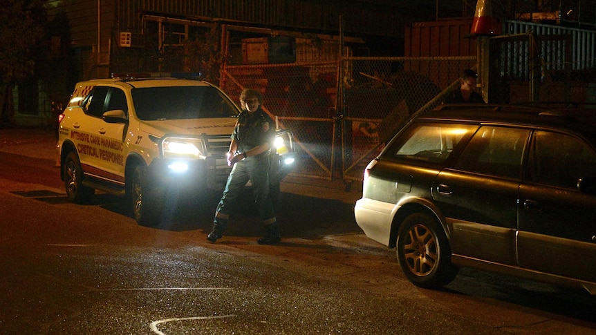 An ambulance officer steps out into the street with a vehicle behind them with the lights on.