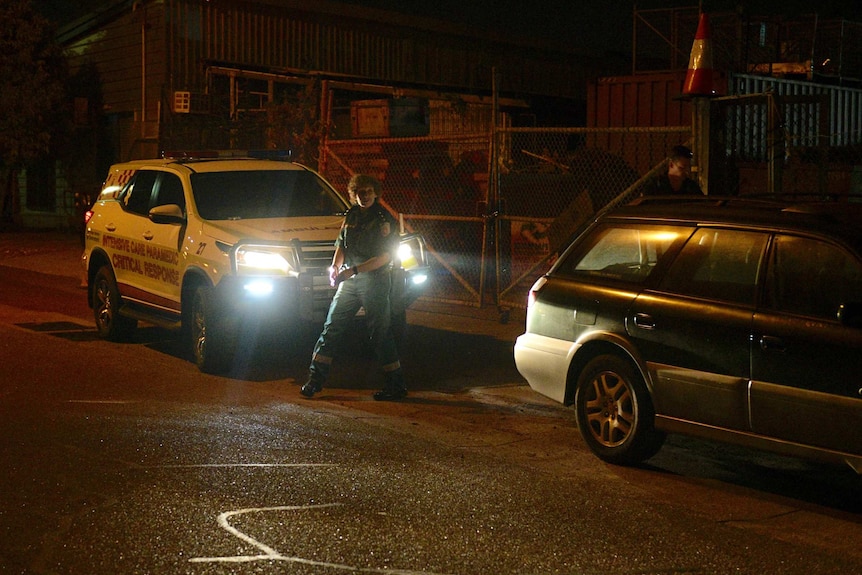 An ambulance officer steps out into the street with a vehicle behind them with the lights on.