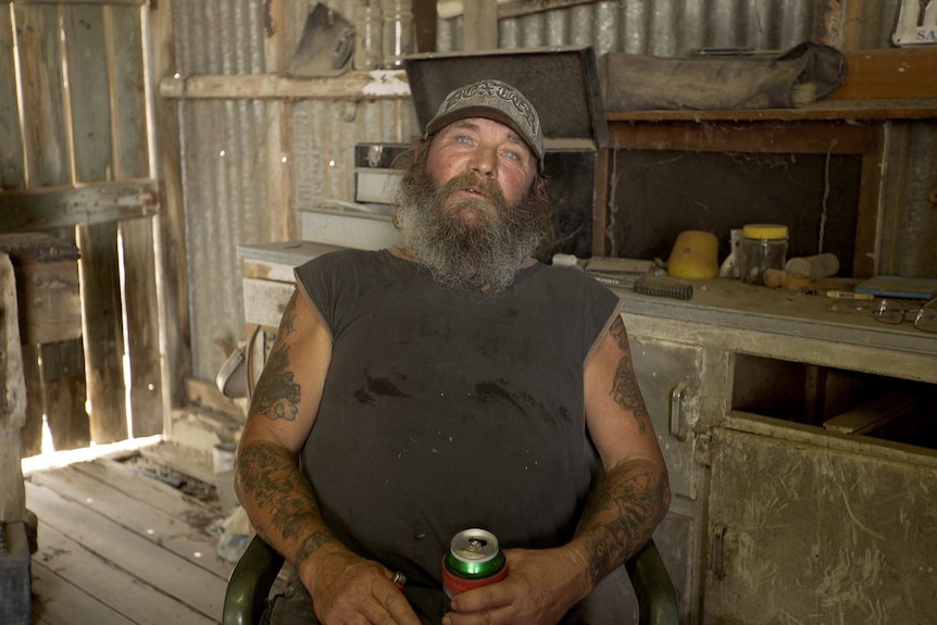 An Australian man leans back in a chair in his shed, surrounded by rustic tools.