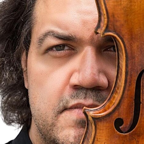 a man's face stares seriously at the camera from behind a violin