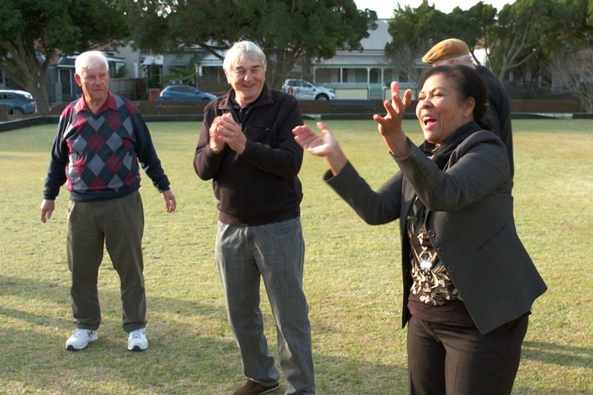 A woman claps her hands and smiles and cheers while standing on a bowling green as two men look on