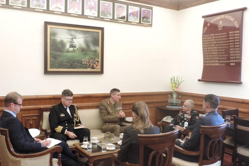 Two middle-aged men in military uniform engage in discussion with other people sitting beside them.