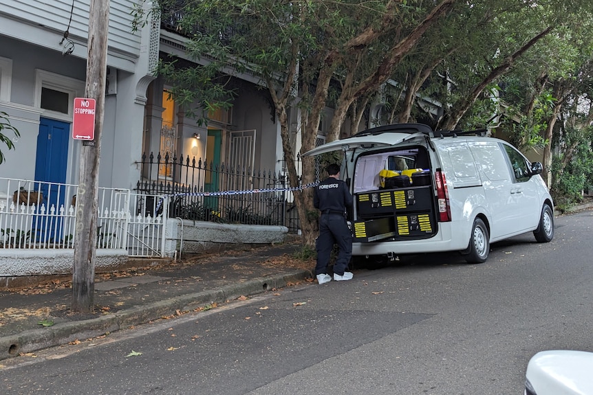 A forensics police officer pulls equipment out from the back of a van that's next to a home with police tape.