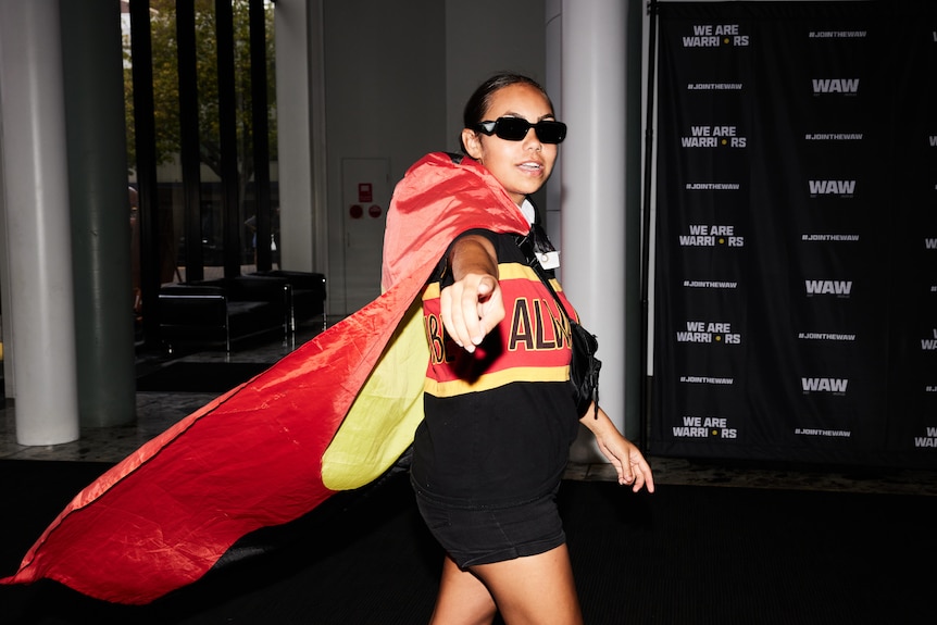 A woman wearing sunglasses and an Aboriginal flag as a cape points at the camera