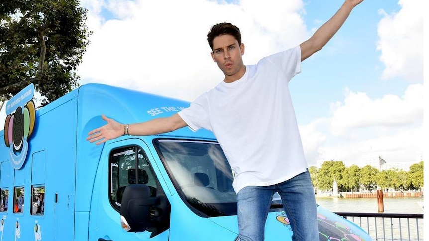 Joey Essex with his hands apart standing in front of a van.