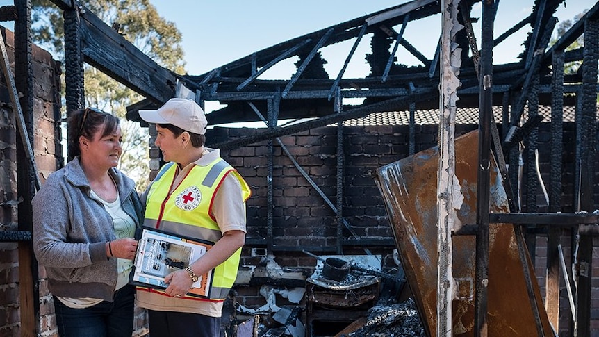 A woman in a grey jacket is comforted by a Red Cross worker in a yellow hi-vis vest as they stand in a burnt out home