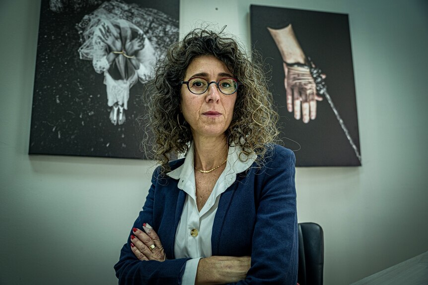 A woman with curly dark hair and glasses sits in an office with her arms crossed, with arty photos of chained hands behind her