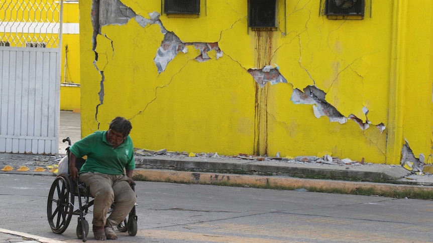 A man sits in a wheelchair in front of a damaged building.