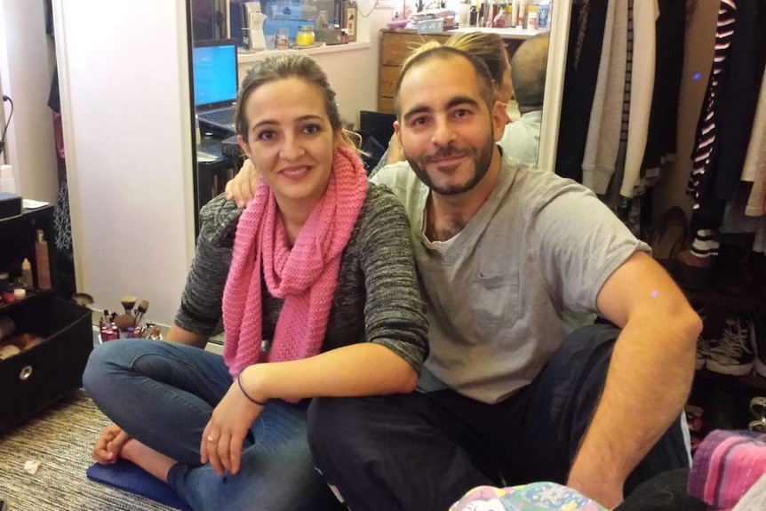 A man and a woman sit on a bedroom floor with his arm around her